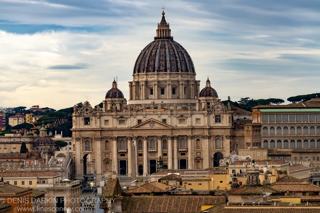Large dome and stone statues above the terracotta rooftops of Rome, soft midday cloud diffusing winter light across the cityscape.