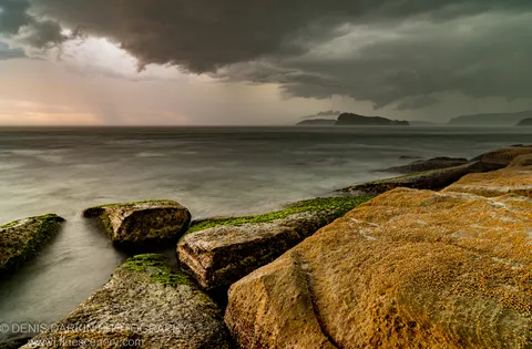 Moss-covered granite boulders anchor the foreground at Umina Beach as a rain curtain falls near distant headlands under heavy cloud.