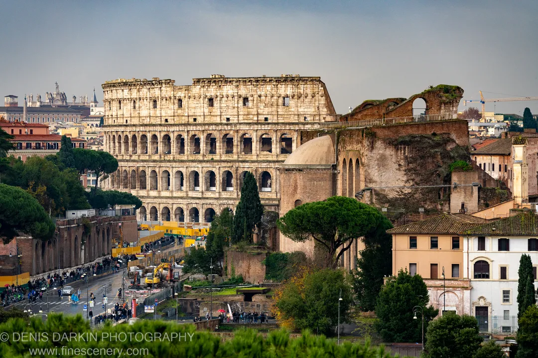 Pale stone arches of the Colosseum rise under a slate-grey sky, framed by Italian pines, with construction barriers visible at the base.