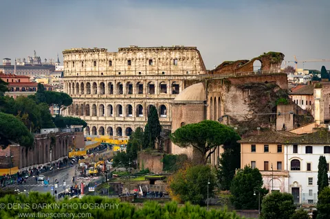 Pale stone arches of the Colosseum rise under a slate-grey sky, framed by Italian pines, with construction barriers visible at the base.