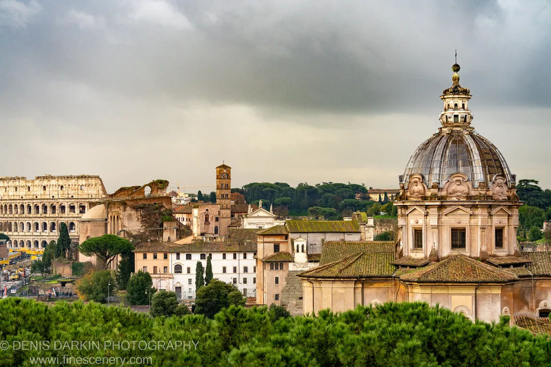 Storm clouds gather above a Rome skyline of ancient stone, terracotta roofs, and a large dome, with green pines filling the foreground.