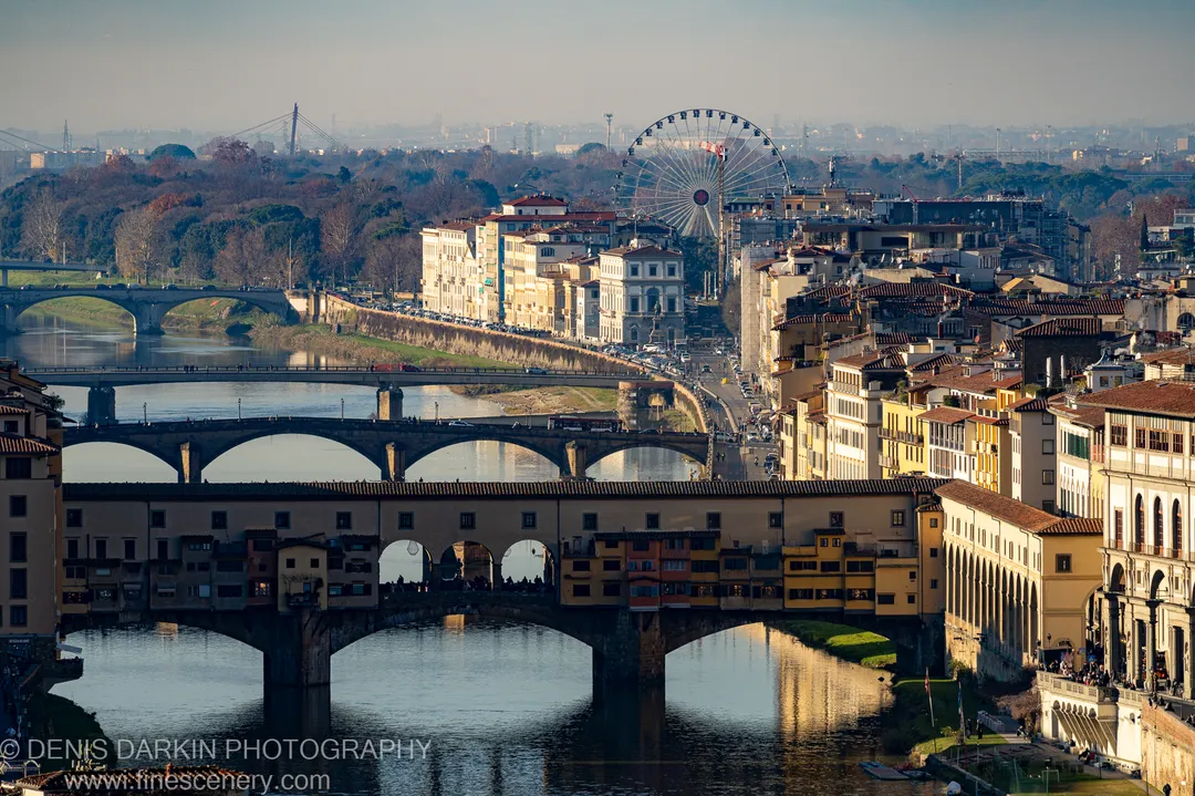 Warm afternoon sunlight falls across ochre facades and terracotta rooftops along a Florence riverbank, with multiple stone bridges layering the foreground.