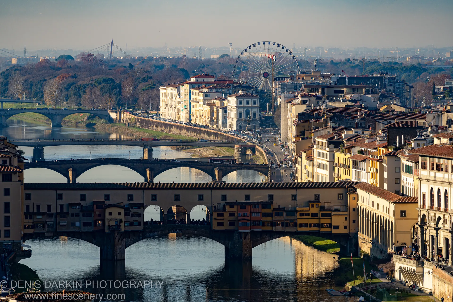 Warm afternoon sunlight falls across ochre facades and terracotta rooftops along a Florence riverbank, with multiple stone bridges layering the foreground.