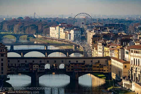 Warm afternoon sunlight falls across ochre facades and terracotta rooftops along a Florence riverbank, with multiple stone bridges layering the foreground.
