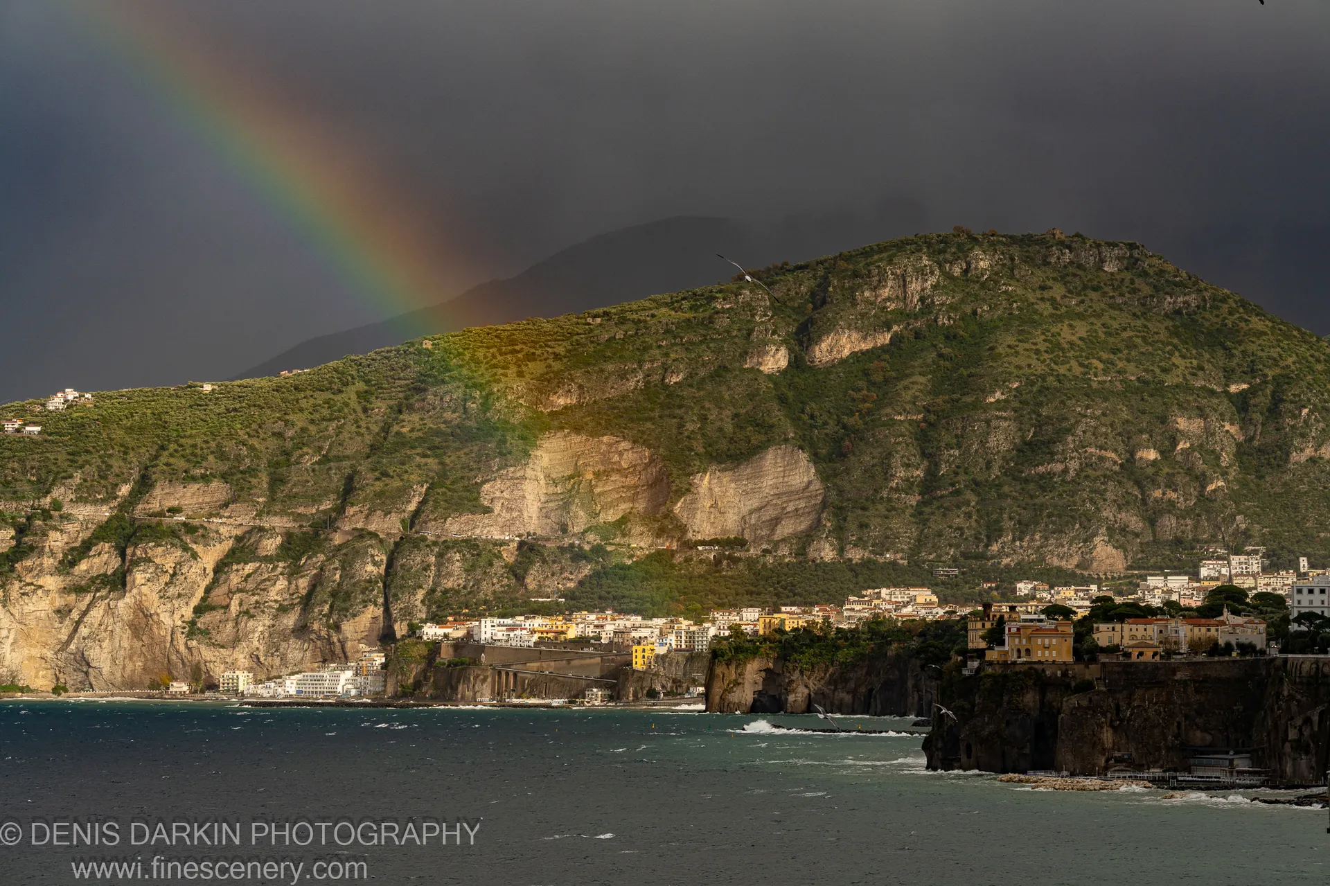 A rainbow arcs over limestone cliffs on the Amalfi Coast, with a coastal village on the steep hillside and whitecaps in the foreground.