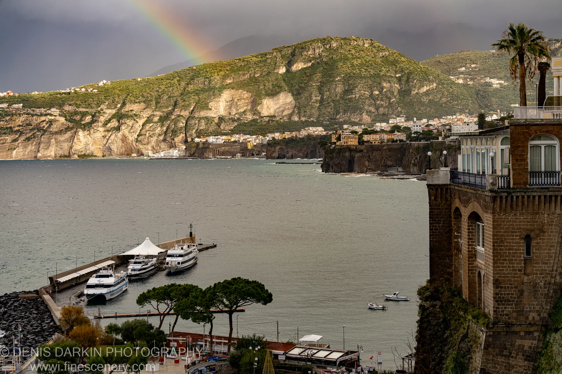 A steep, forested mountain towers over the Amalfi harbor, a rainbow arching through storm clouds above while white boats rest in grey water.