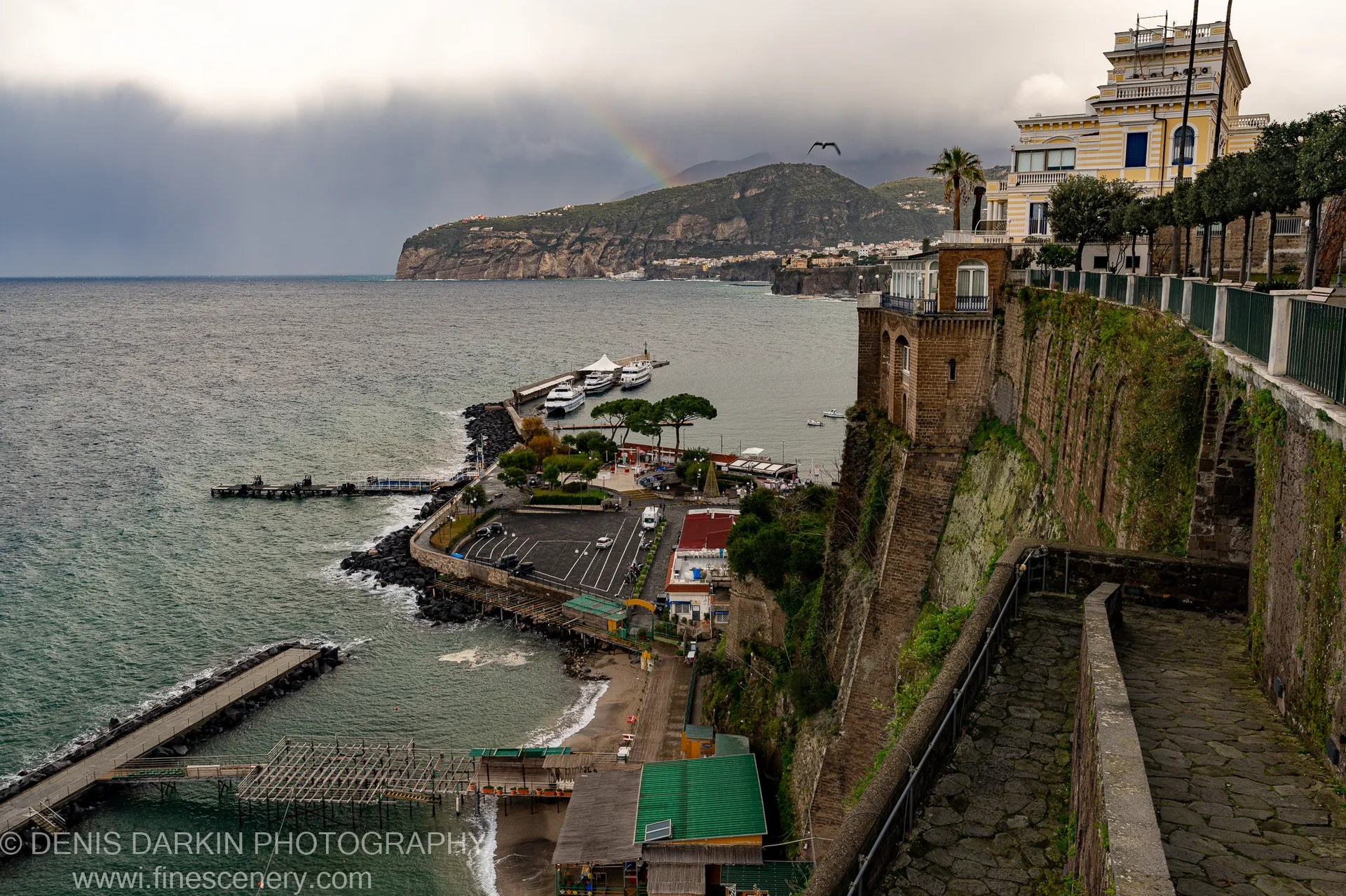 Moss-covered stone retaining walls descend toward an Amalfi harbor, a yellow building perched at the cliff edge above.