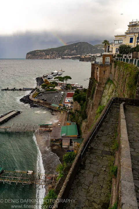 A cobblestone walkway leads toward an Amalfi harbor overlook, storm clouds and a faint rainbow above the headland in the distance.