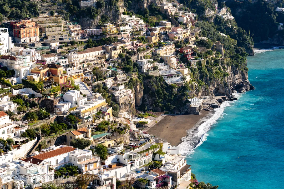 Terraced stone buildings cling to a steep limestone cliff above turquoise water on the Amalfi Coast, a black sand beach below.