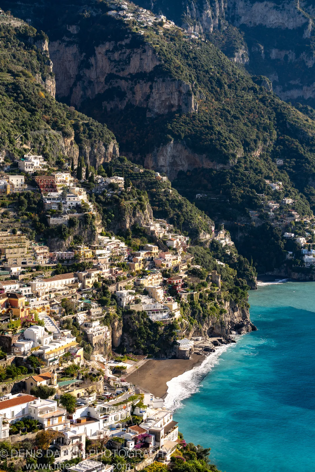 Terraced facades on rugged cliffs above a dark sand beach at Sorrento, with turquoise water breaking at the base in winter morning light.