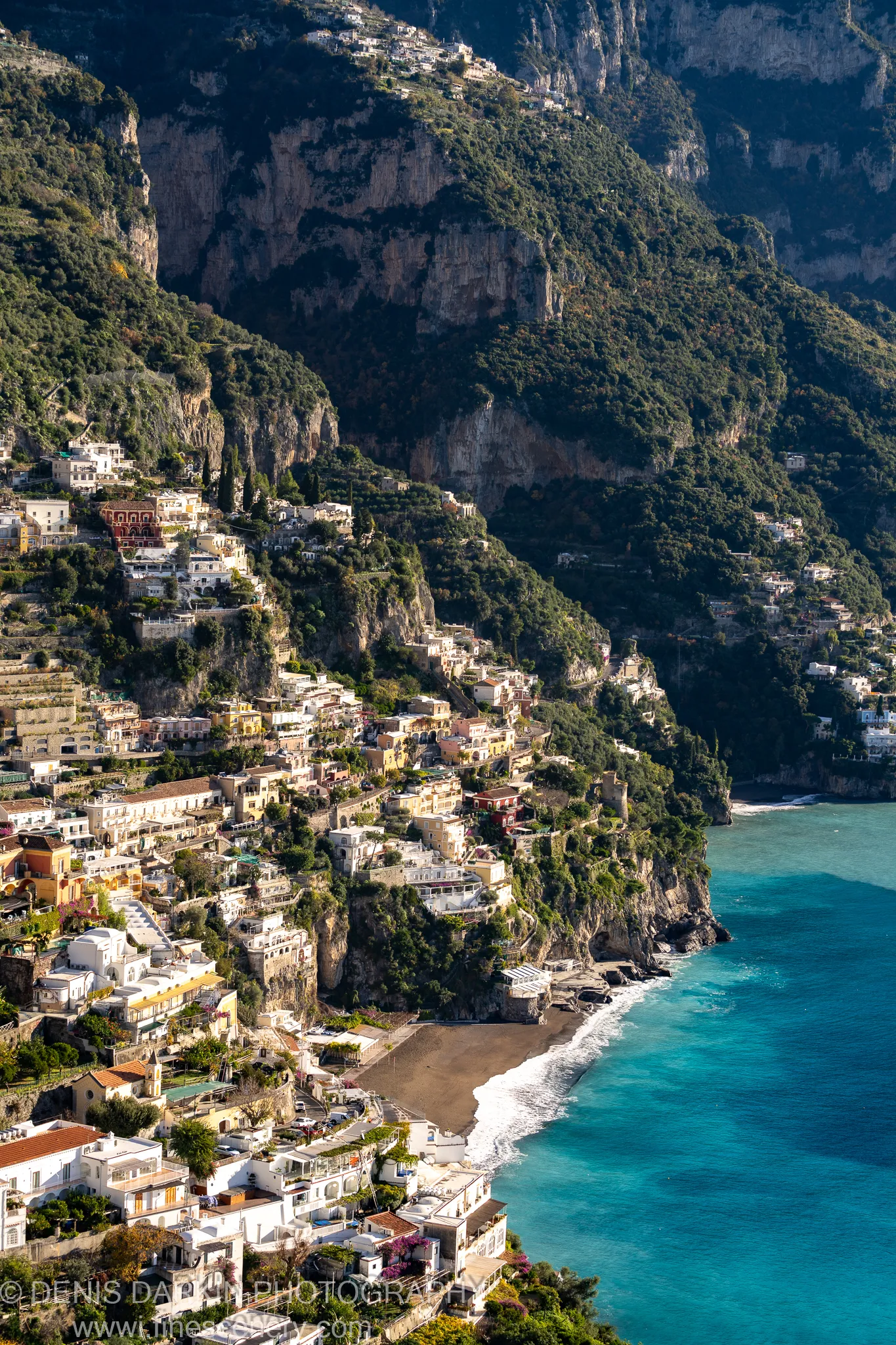 Terraced facades on rugged cliffs above a dark sand beach at Sorrento, with turquoise water breaking at the base in winter morning light.