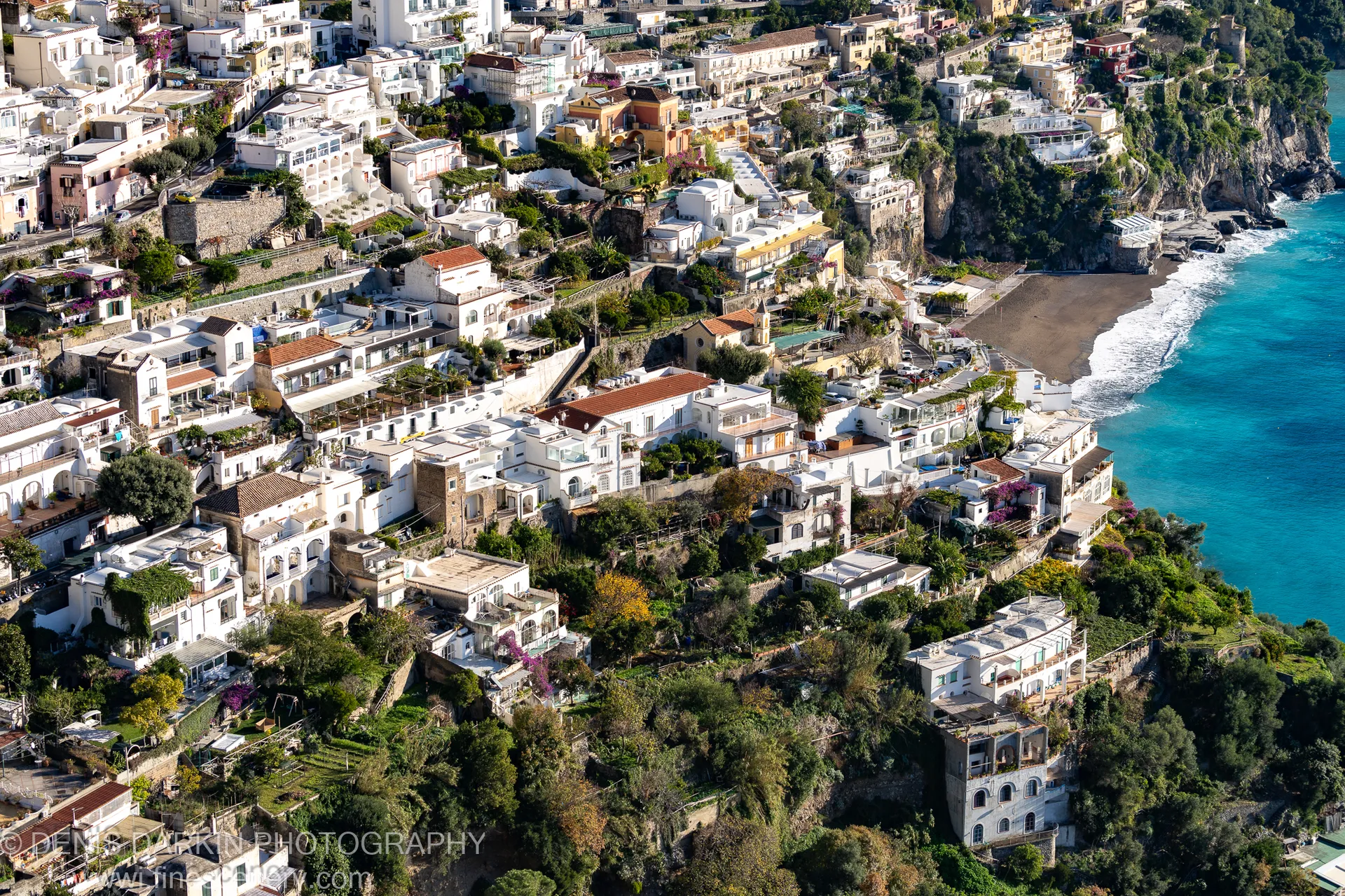Stone walls and bare branches in shadow overlook an electric blue bay and dark sand beach at the Sorrento coast, December 2024.