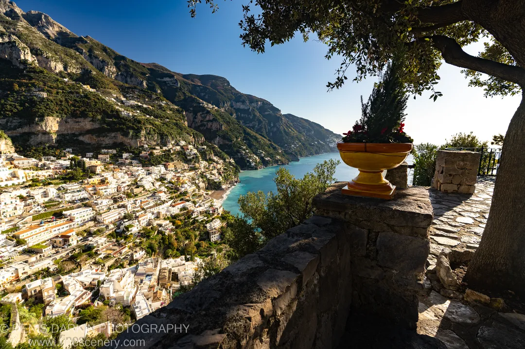 A weathered stone wall and yellow ceramic planter sit in shadow before the sunlit white buildings on green slopes above the bay at Sorrento.