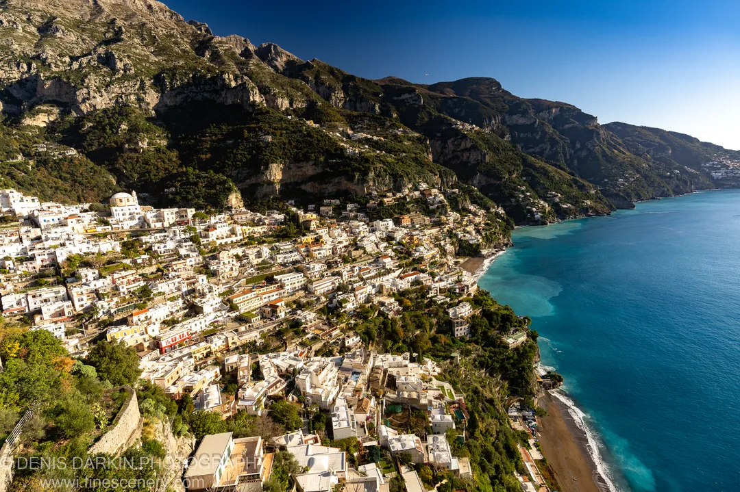Terraced whitewashed buildings on rugged mountain slopes above the azure bay at Sorrento, with deep shadow in the cliffside ravines, December morning light.
