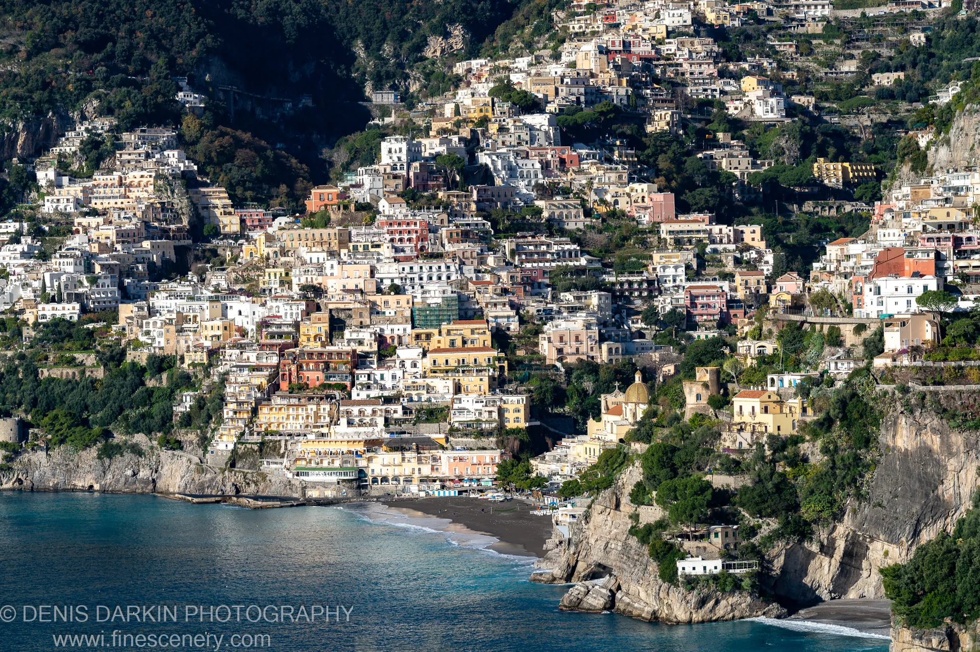 Pastel buildings on a steep cliffside above a black-sand beach at Sorrento, with a golden church dome visible among the tightly packed terraces.
