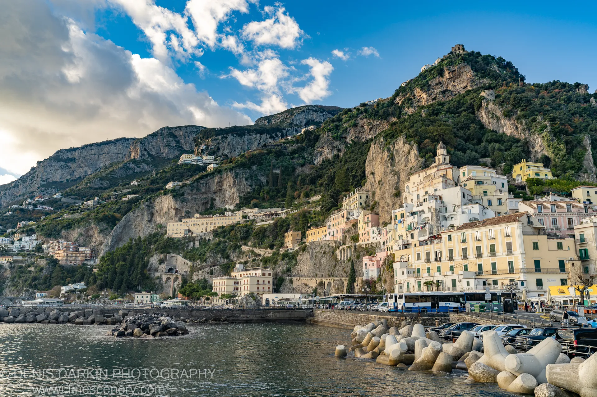 Pastel buildings above a harbor lined with concrete tetrapods at Sorrento, a mountain-top ruin visible at the cliff summit in midday sun.