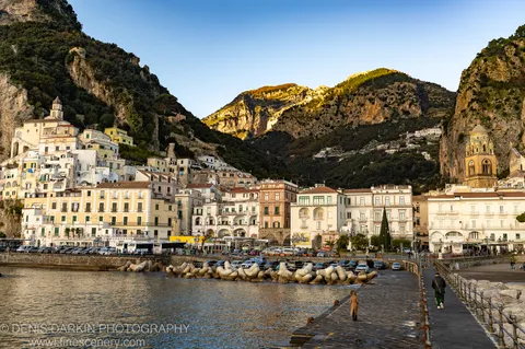 Mountain peaks in low winter sun above Sorrento's harbor at midday, stone promenade and concrete breakwaters in the foreground, lone figures in shadow.