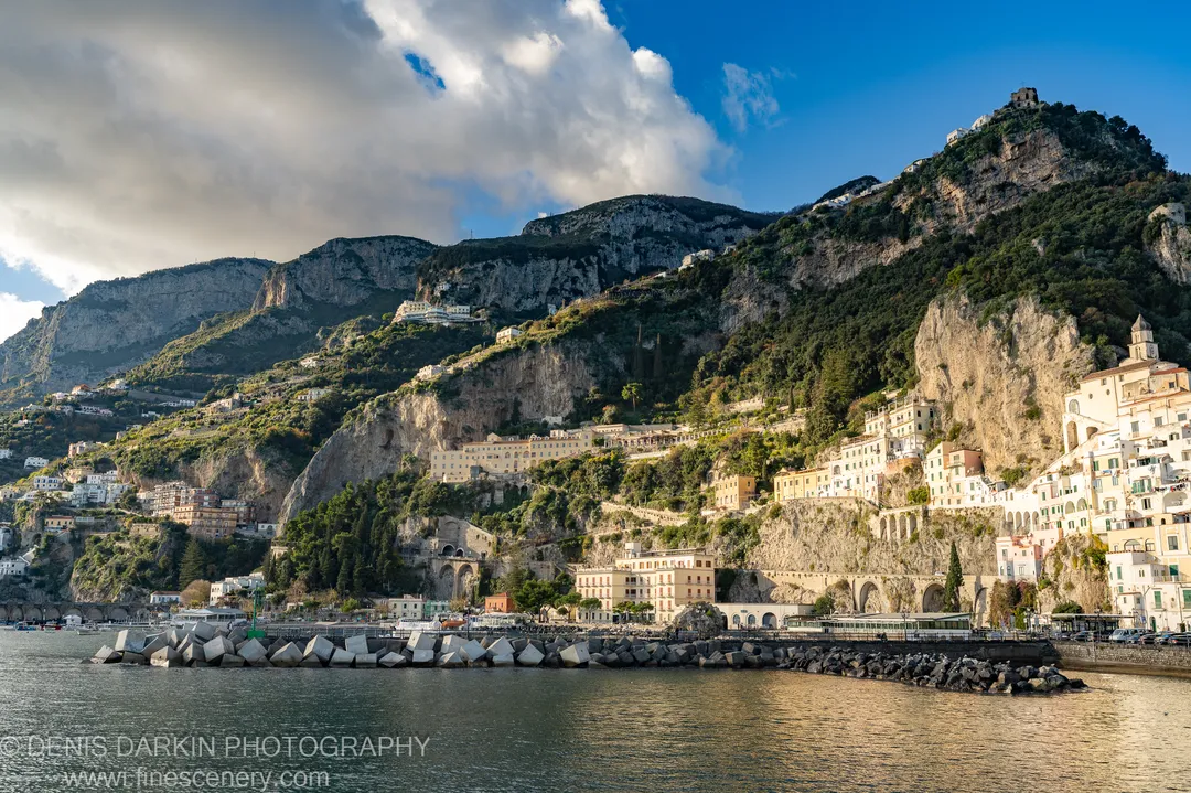 Whitewashed buildings on limestone cliffs above concrete tetrapods at Sorrento, large clouds over the mountain peaks in afternoon light, December 2024.