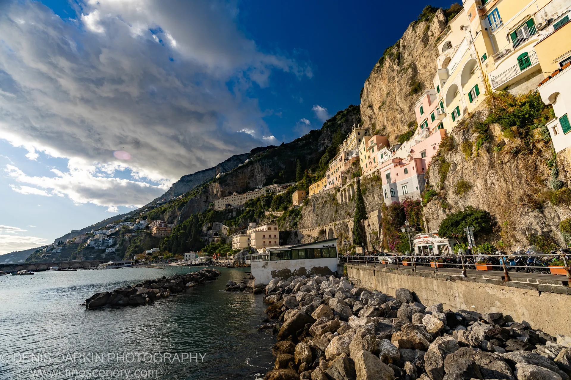 Pastel houses on a steep cliffside above a calm harbor on the Amalfi Coast, with dark volcanic rocks in the foreground and heavy cloud overhead.