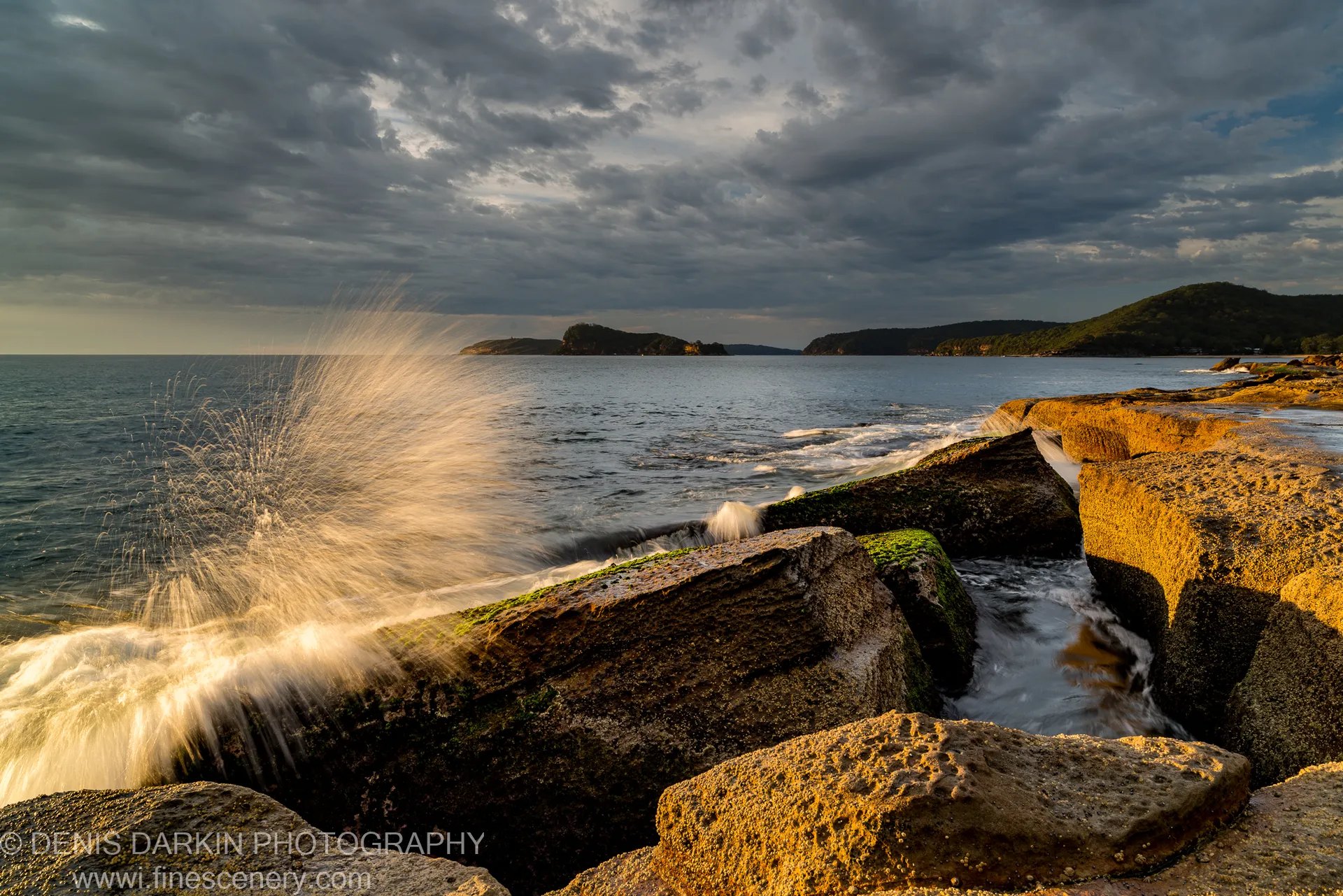 A wave crashes against mossy sandstone rocks at Umina Beach at dawn, golden spray catching the first light below a stormy sky.