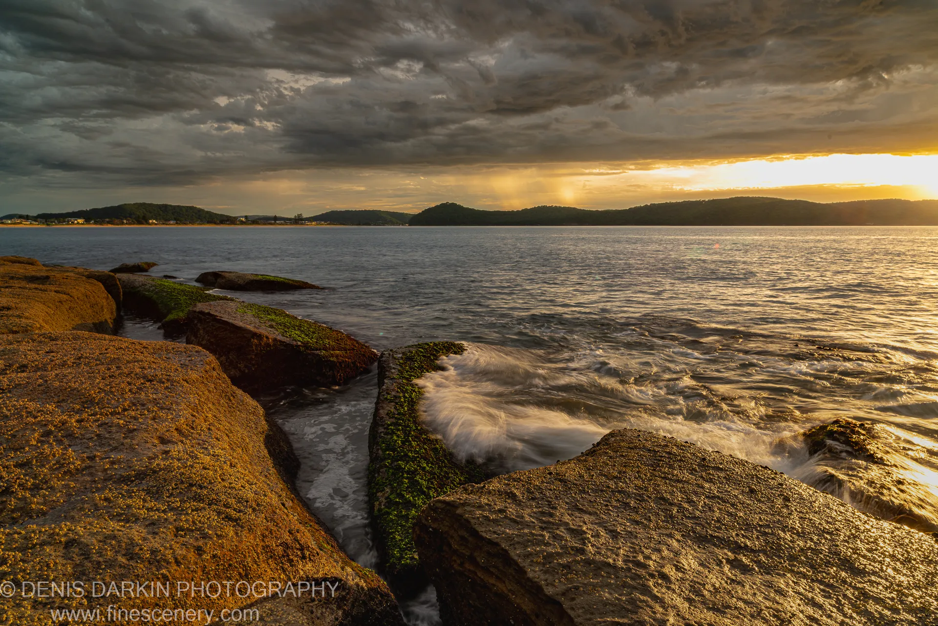 Golden morning sunlight breaks through storm clouds over the bay at Umina Beach, with barnacle-covered foreground rocks and distant hills visible.