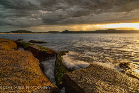 Golden morning sunlight breaks through storm clouds over the bay at Umina Beach, with barnacle-covered foreground rocks and distant hills visible.