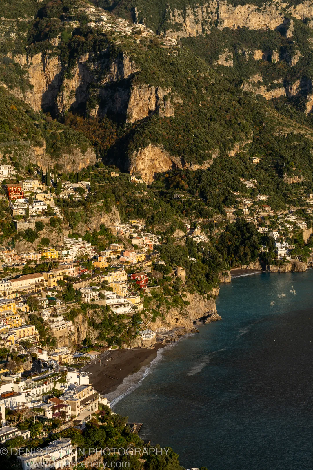 Warm late-afternoon light falls on terraced limestone cliffs and pastel buildings overlooking a dark sand beach on the Amalfi Coast.