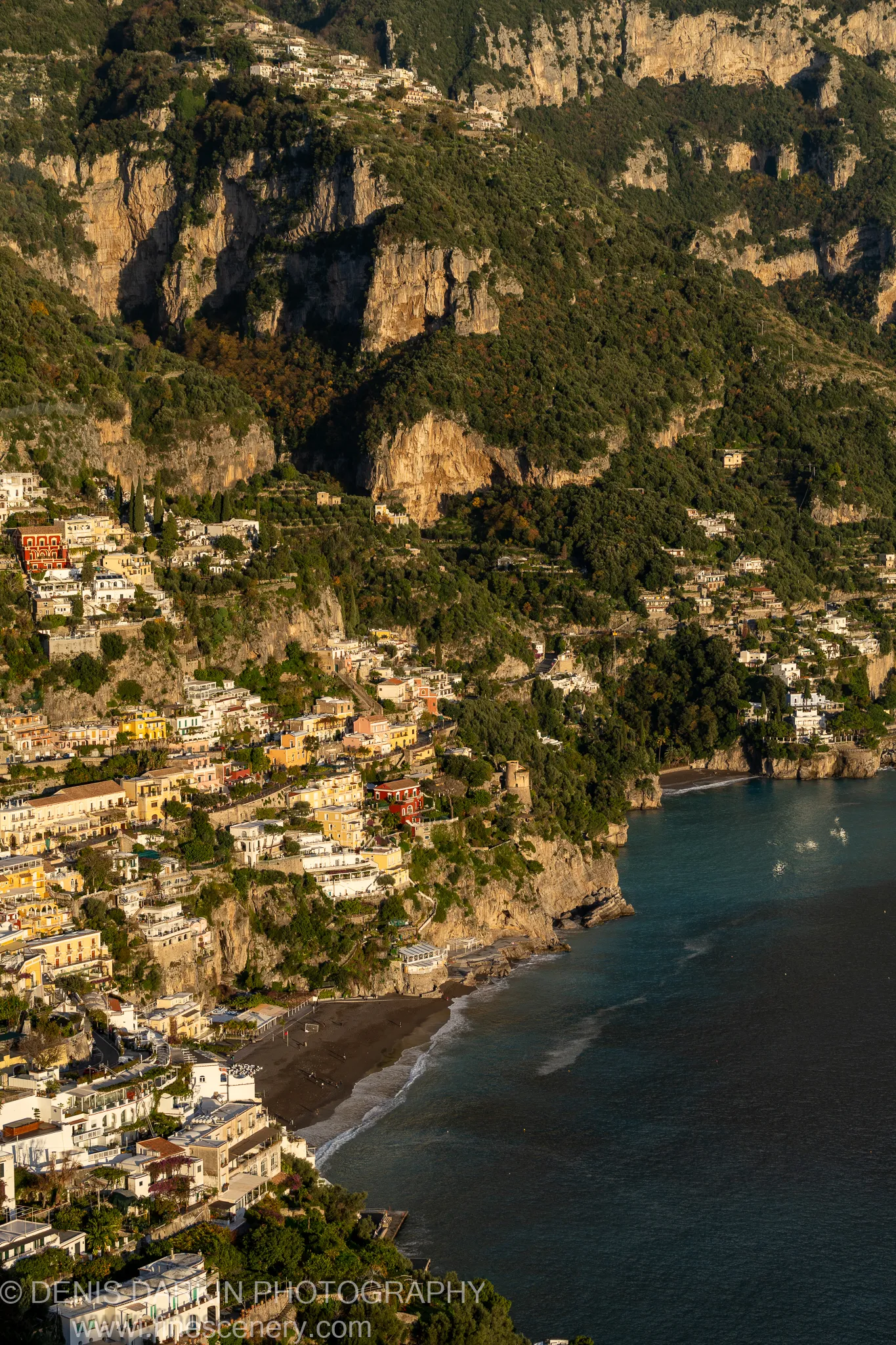 Warm late-afternoon light falls on terraced limestone cliffs and pastel buildings overlooking a dark sand beach on the Amalfi Coast.