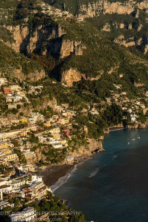 Warm late-afternoon light falls on terraced limestone cliffs and pastel buildings overlooking a dark sand beach on the Amalfi Coast.