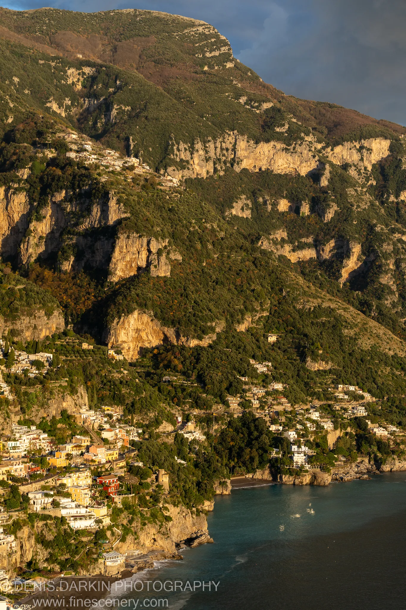 Sunlight strikes the rugged limestone face of a steep mountain, casting shadows across terraced buildings on the Amalfi Coast at golden hour.