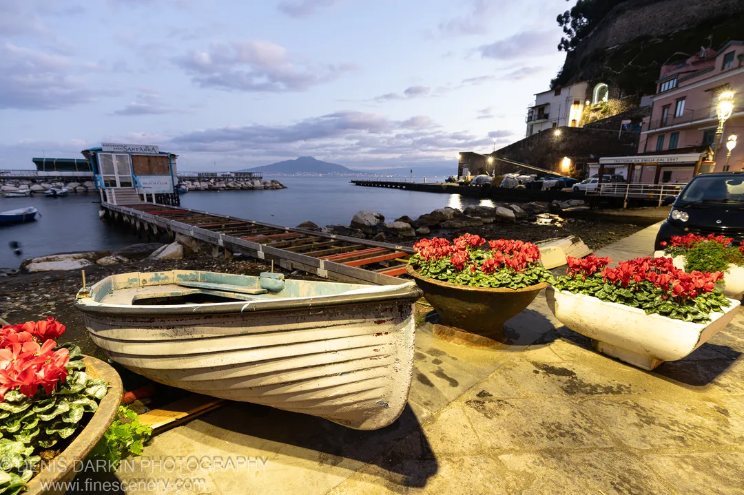 A white rowboat rests on a stone quay beside red cyclamen planters, the wooden pier and mountain silhouette visible in the Amalfi Coast dawn beyond.