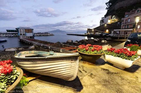 A white rowboat rests on a stone quay beside red cyclamen planters, the wooden pier and mountain silhouette visible in the Amalfi Coast dawn beyond.