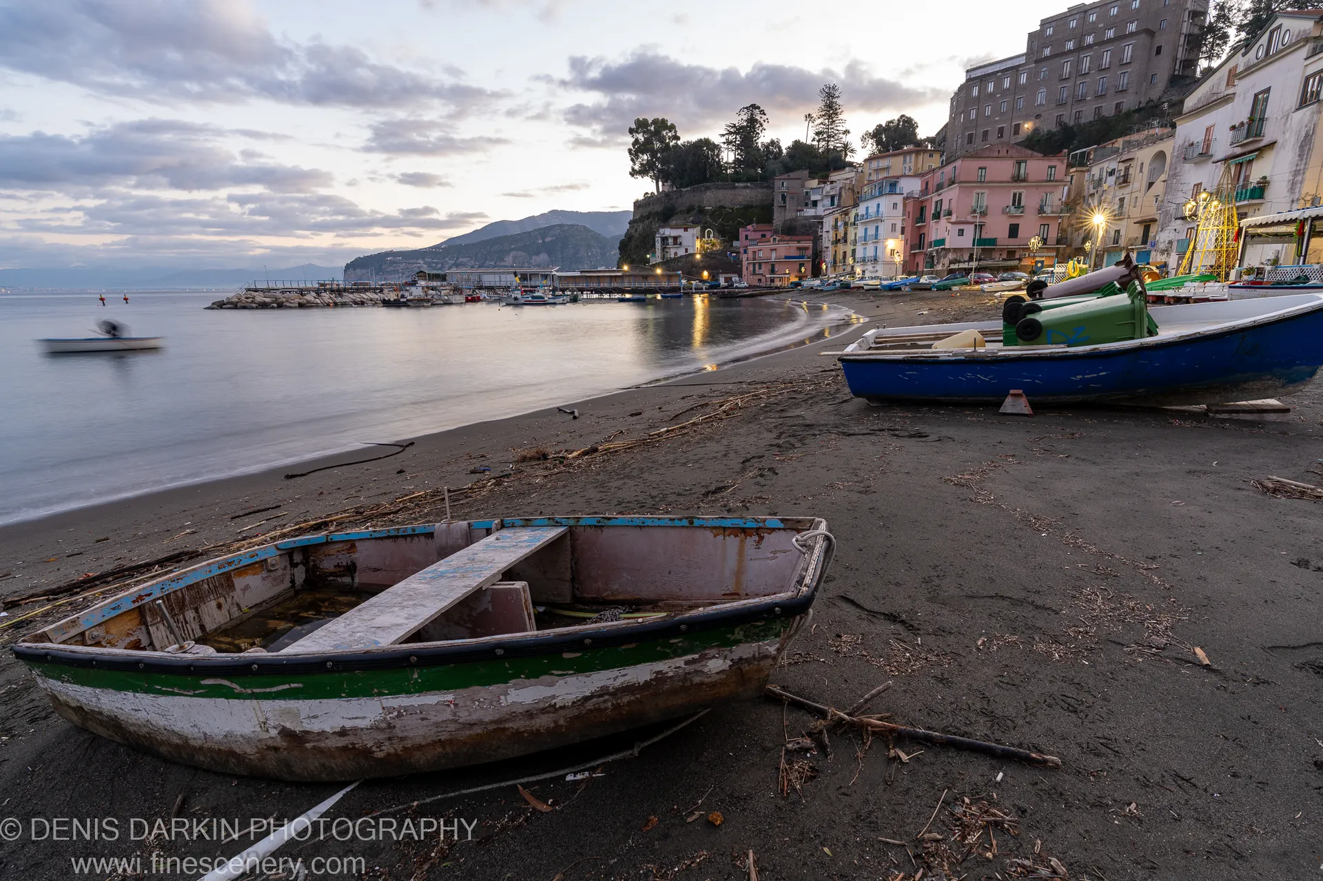 Weathered boats with peeling paint rest on dark volcanic sand below colorful buildings lit by streetlights at Amalfi Coast dawn.