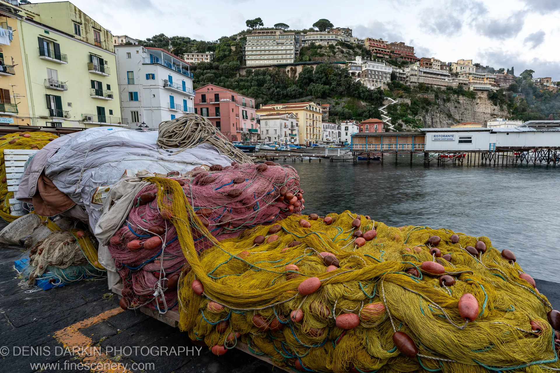 Yellow and pink fishing nets rest on a stone quay at dawn on the Amalfi Coast, the hillside town visible beyond the calm harbor.
