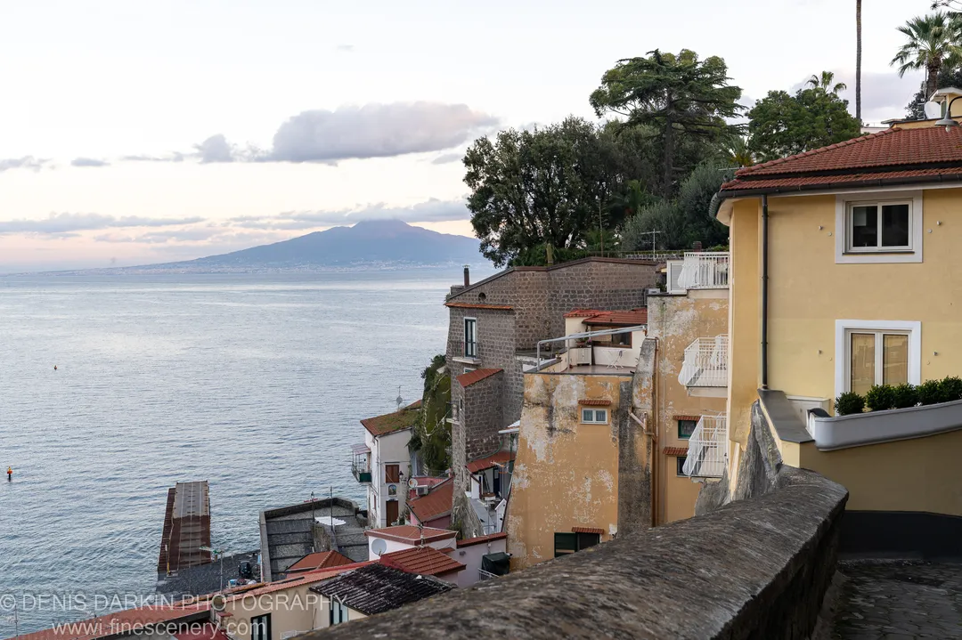 A stone wall borders a calm Amalfi Coast bay at dawn, with ochre buildings on the hillside and a mountain silhouette in the distance.