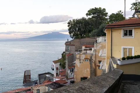 A stone wall borders a calm Amalfi Coast bay at dawn, with ochre buildings on the hillside and a mountain silhouette in the distance.