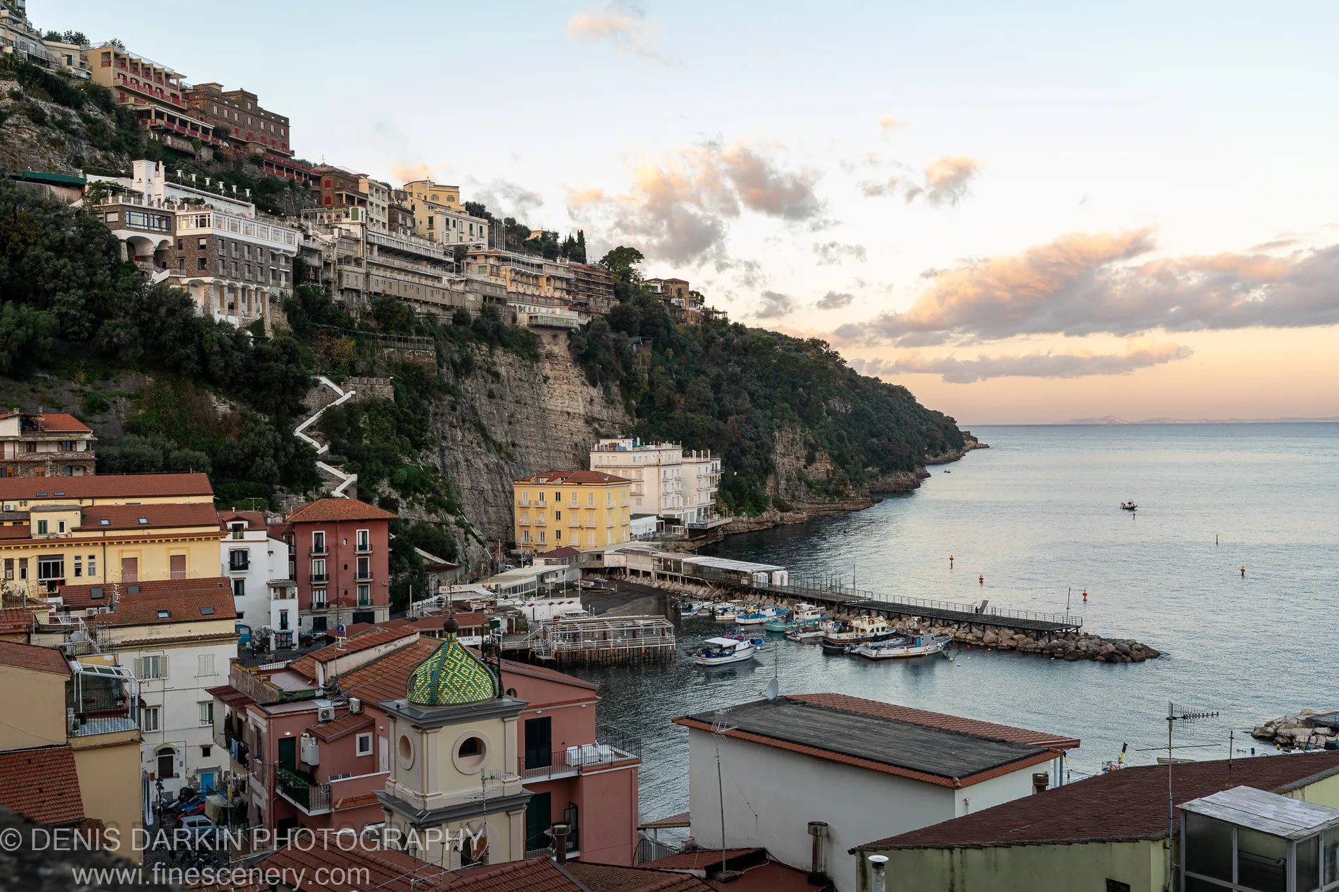 Terracotta-roofed buildings step down to a sheltered harbor pier on the Amalfi Coast, the church dome prominent in soft December dawn light.