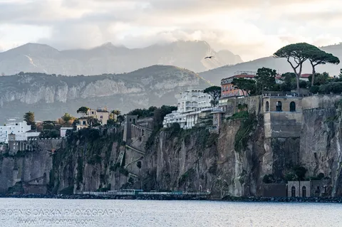 A steep limestone cliff rises from calm water, topped with white villas and stone arches, umbrella pines on the crest and misty mountains beyond.