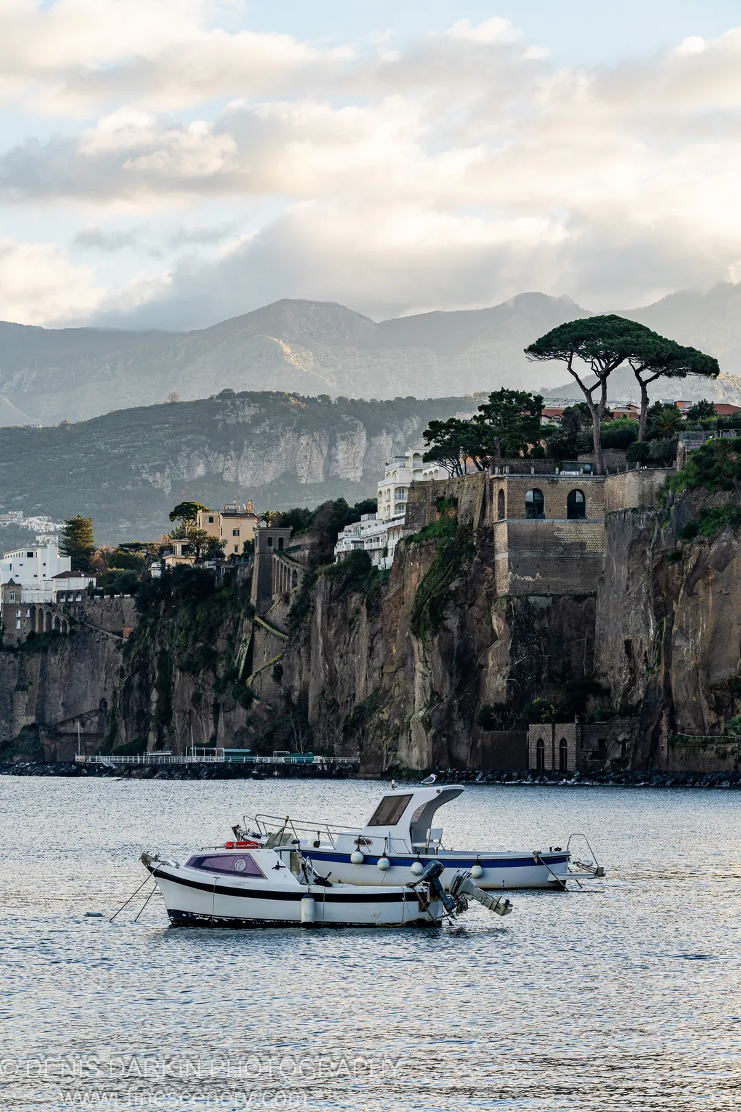 Two small boats on rippling water, white villas in the cliff, umbrella pines against a pale winter sky. Amalfi Coast.