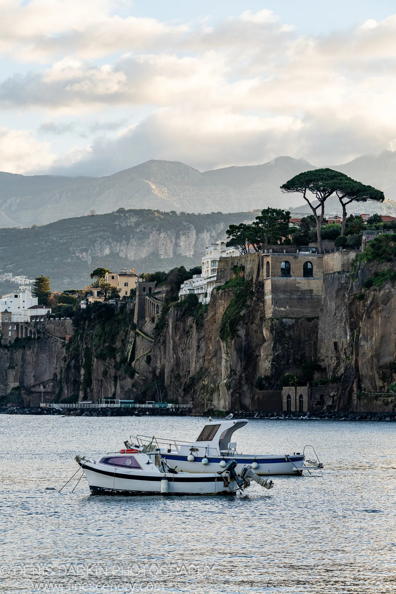Two small boats on rippling water, white villas in the cliff, umbrella pines against a pale winter sky. Amalfi Coast.