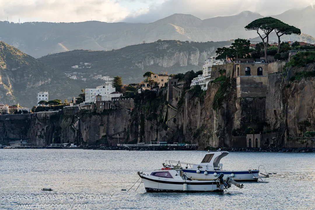 Two boats moored on calm water, rugged cliffs with white buildings at their edge above, soft light on distant mountain slopes. Amalfi Coast.