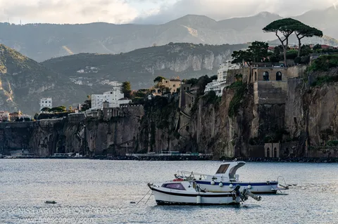 Two boats moored on calm water, rugged cliffs with white buildings at their edge above, soft light on distant mountain slopes. Amalfi Coast.