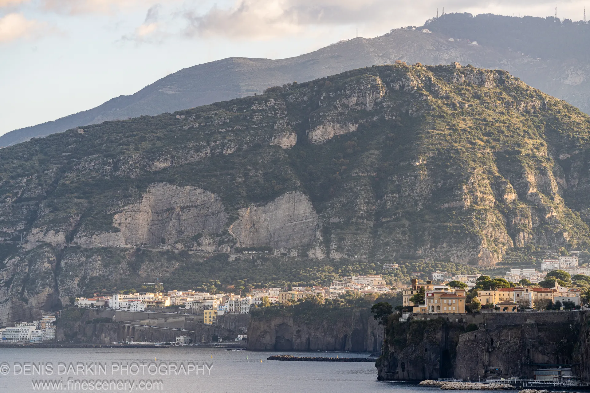 Warm light on the facades of an Amalfi Coast hillside village at dawn, the calm sea below in cool pale tones. December 2024.