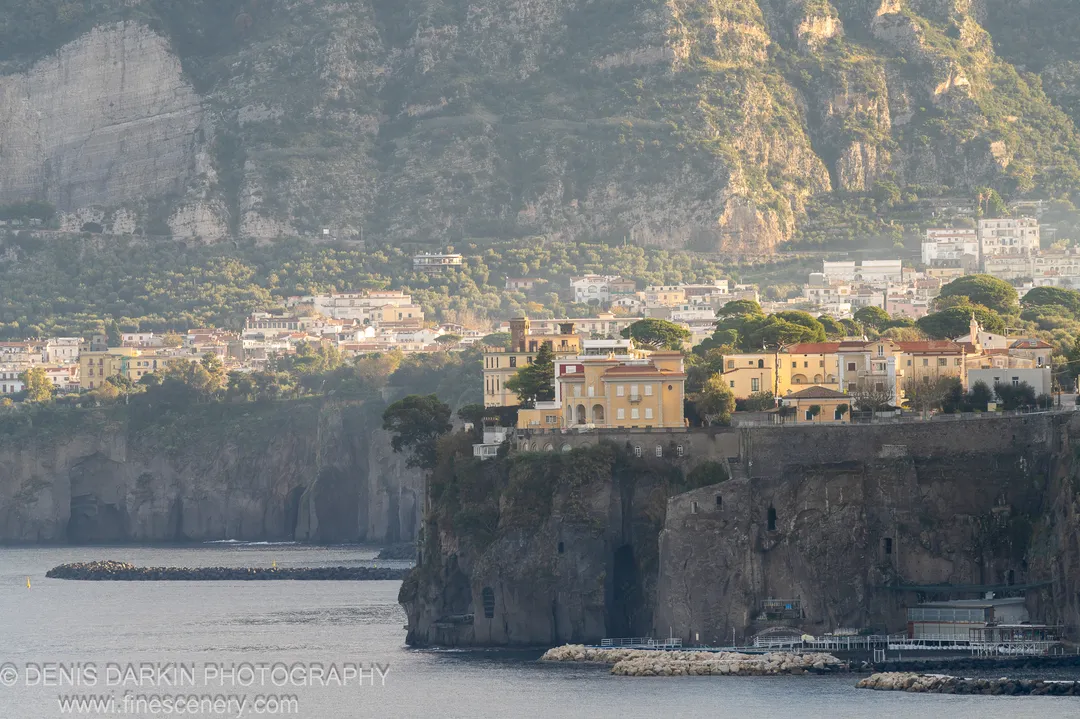 Sunlit upper cliff above a shadowed Amalfi Coast harbor, warm light on the rock face and cool water below. December 2024.