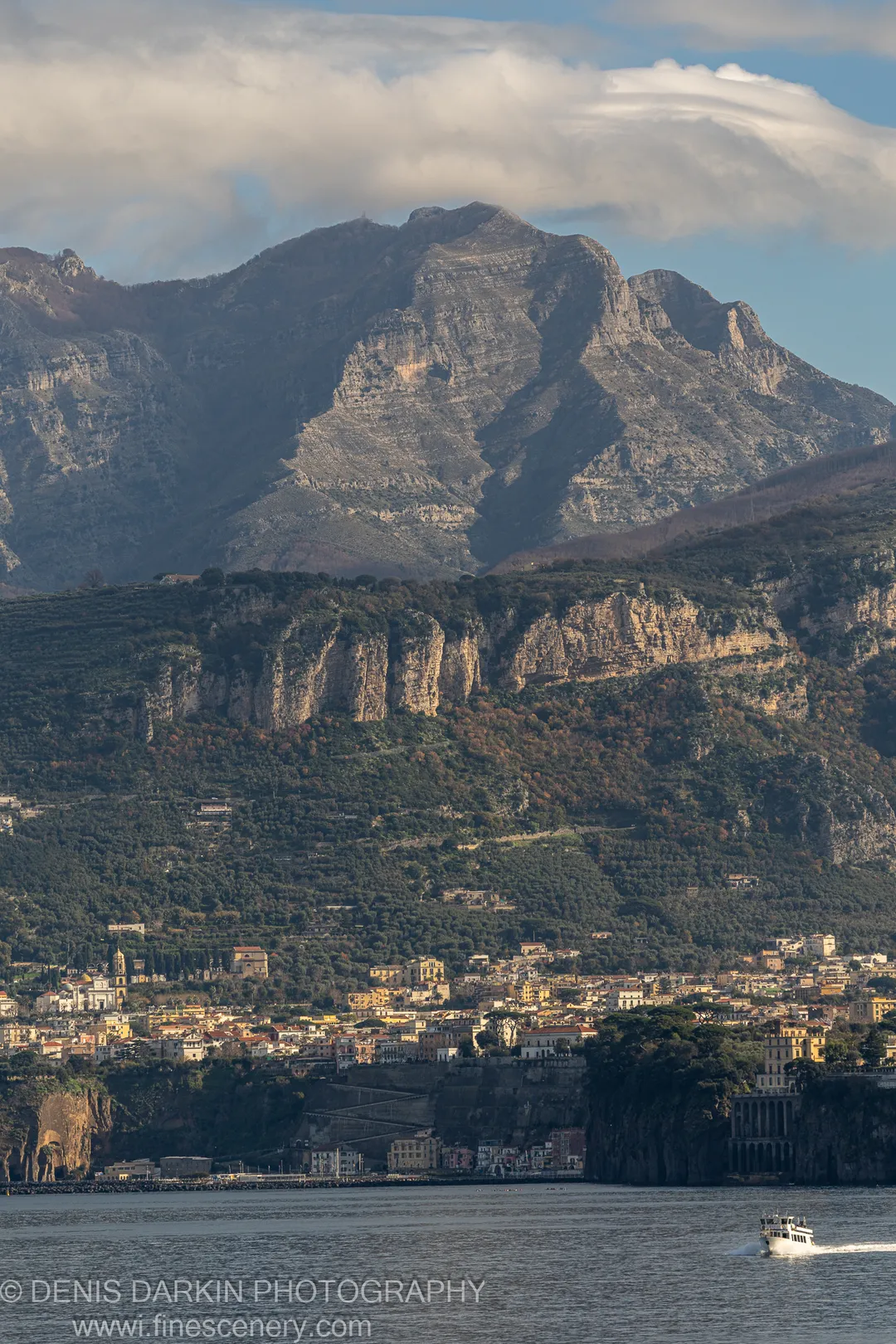 Compact coastal settlement beneath terraced hillsides and a flat shelf cloud over the summit, Amalfi Coast.