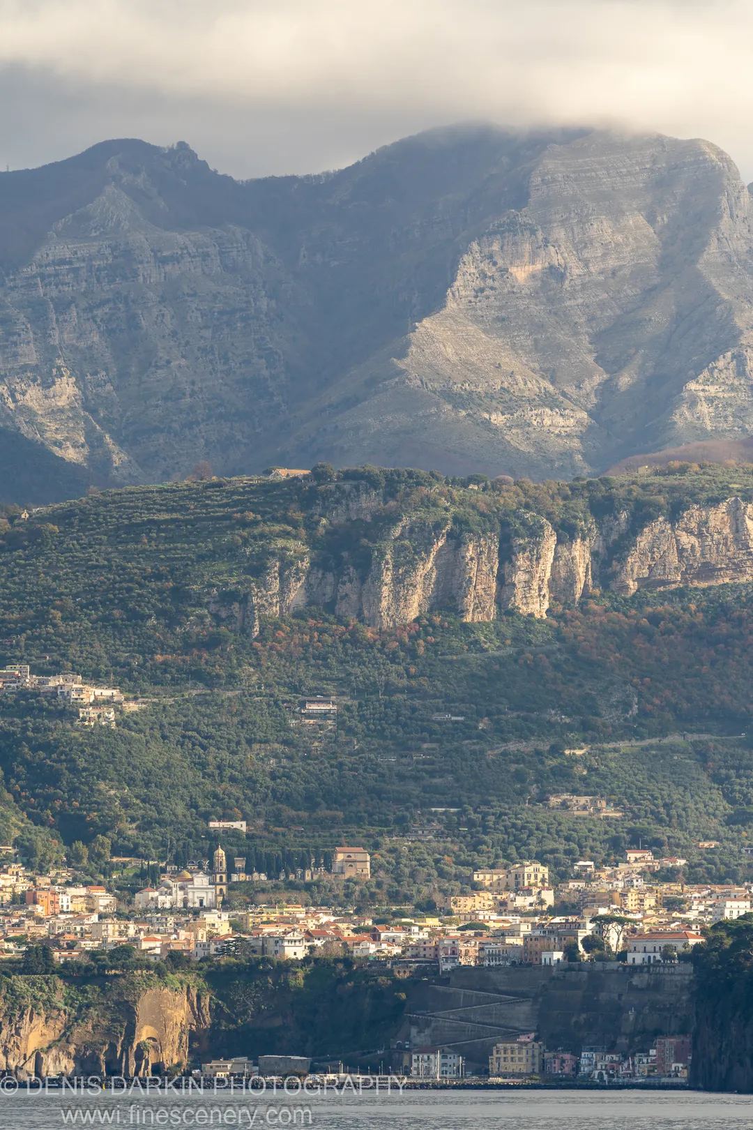 Dense coastal town against a sheer limestone cliff rising to mist-shrouded peaks, a stone fortification in the foreground below. Amalfi Coast.
