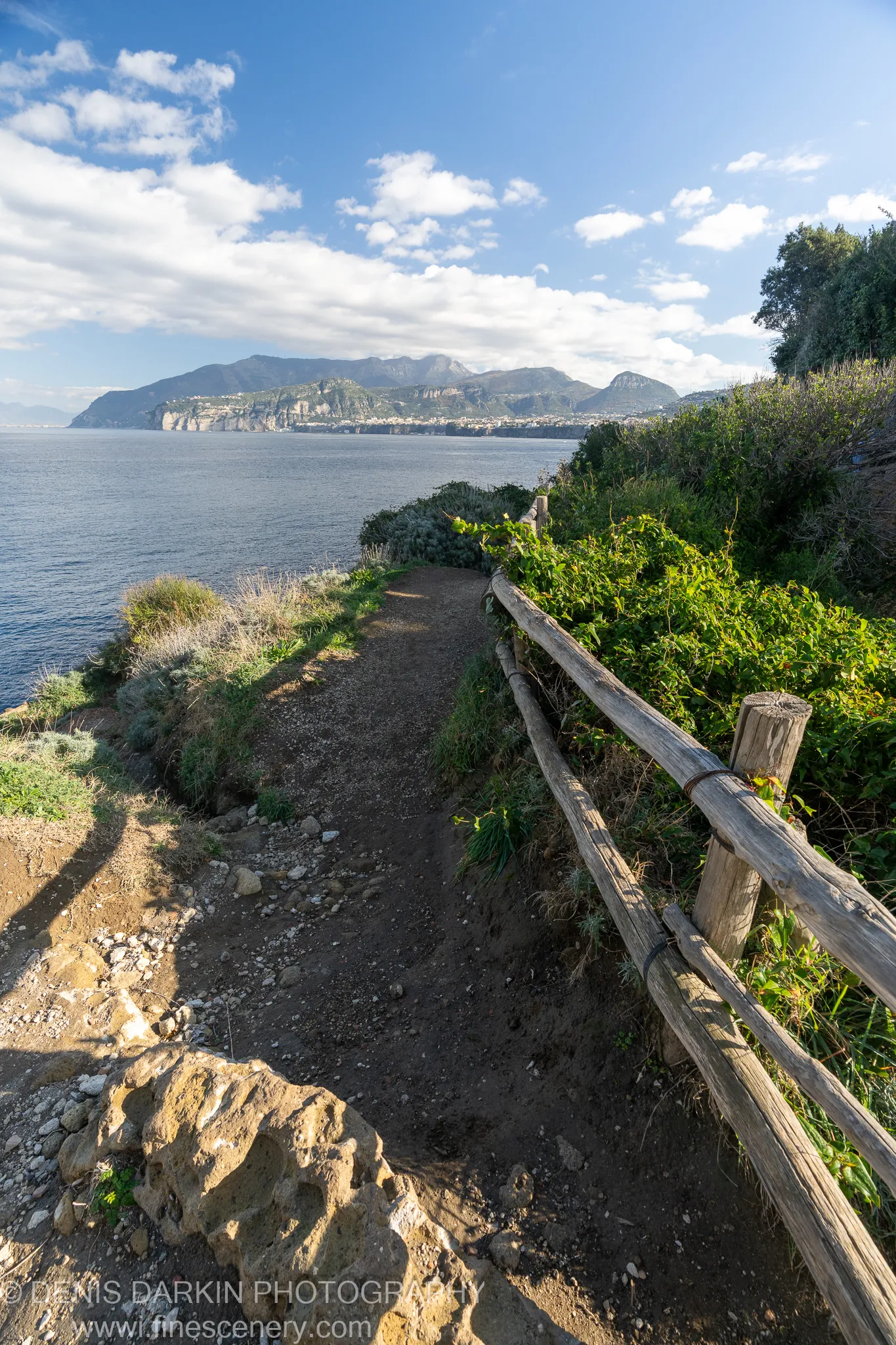 Dirt cliff-edge path with a weathered wooden fence, textured limestone in the foreground, distant Amalfi Coast mountains beyond.