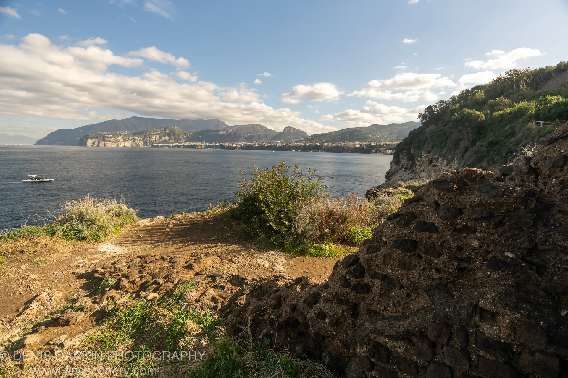 Dark volcanic stone at the cliff edge above a sun-drenched bay, deep blue water extending to distant mountains, Amalfi Coast.