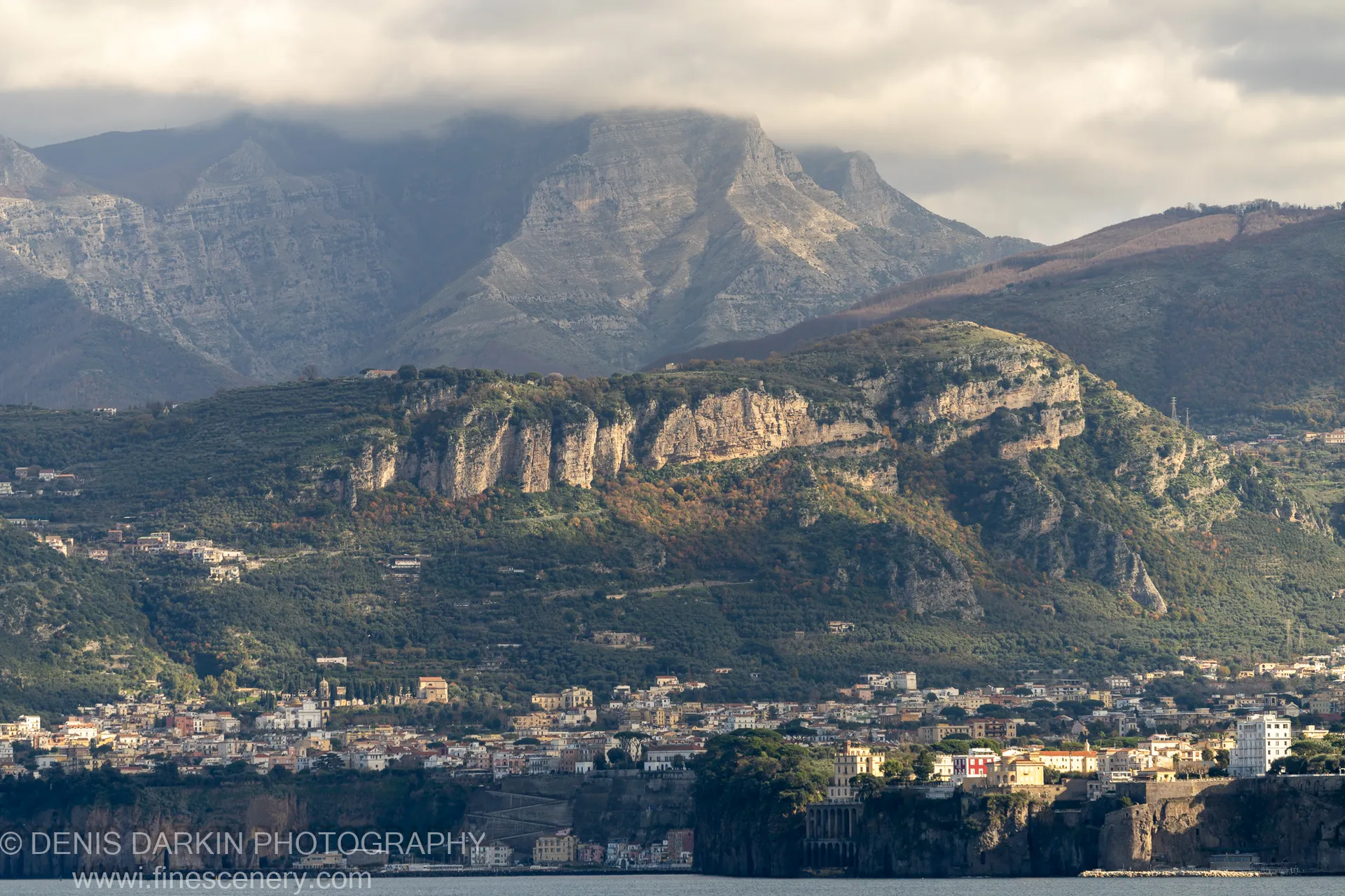 Massive stratified mountains over an Amalfi Coast town on steep cliffs, low clouds at the peaks, terracotta roofs in the foreground.