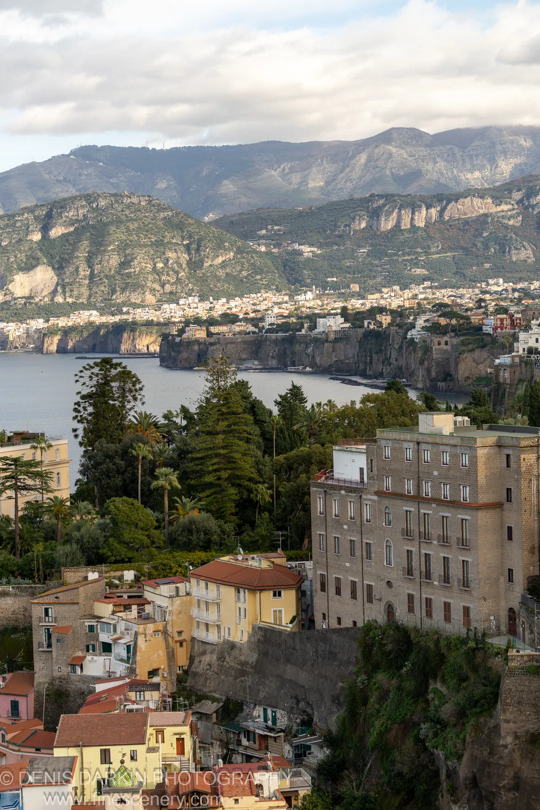 Terracotta-roofed buildings on a steep Amalfi Coast slope, stone walls in soft directional light, a calm bay and distant mountains beyond. December 2024.